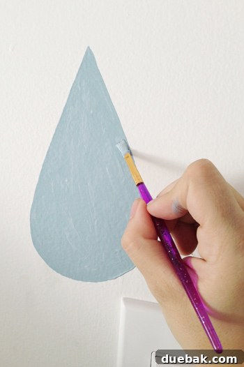 Hand Painting Blue Raindrop On Wall Mural With Small Brush - A hand carefully painting a blue raindrop onto a wall mural using a small craft brush.