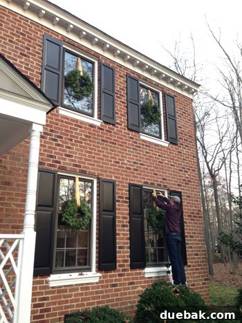 Effortless Window Wreath Styling 9 A Christmas wreath being meticulously hung on a downstairs window from the exterior using a sturdy ladder, demonstrating the adapted installation method required for different window types.