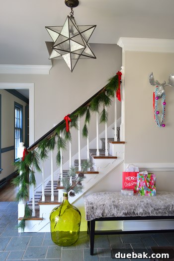 Staircase adorned with faux garland and red ribbons, leading to a cozy foyer bench with a furry blanket.