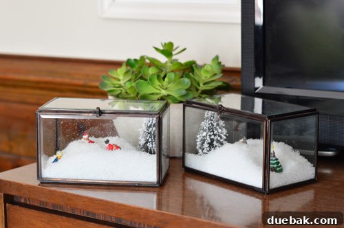 Close-up of crafting supplies: two glass boxes, a bag of fake snow, mini trees, and a package of tiny ceramic dogs.