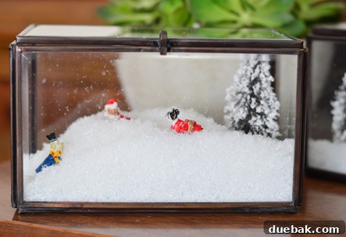 Two completed snow-filled glass boxes with mini trees and ceramic dogs displayed on a media cabinet.