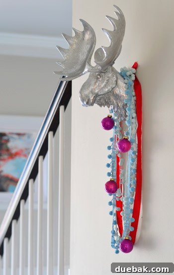 A silver moose head adorned with festive ribbons and pink ornaments, hanging on a blue wall.