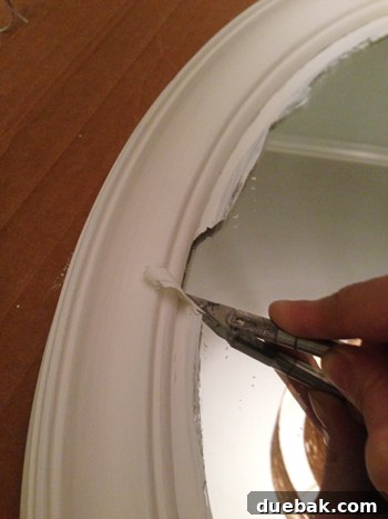 A close-up of a hand using an exacto knife to scrape paint off a mirror's surface, showing the clean separation.