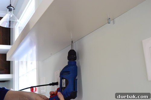 Using a power drill to quickly and securely screw an IKEA floating shelf to its wall bracket, illustrating an efficient installation technique.