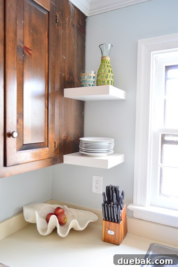 Two short IKEA floating shelves placed next to a kitchen window, beautifully holding a stack of white plates and a stylish carafe, blending utility with charm.