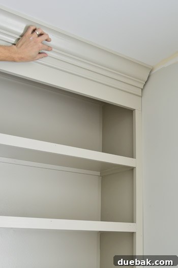 Craft Your Own Library Haven 16 A hand holding a piece of decorative crown molding in place on top of a DIY bookshelf, illustrating the final aesthetic touch.