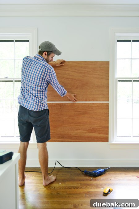 dry fitting plywood planks on wall between two windows to become base for gluing cork board tiles to create cork board wall
