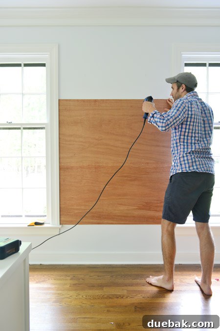 using power drill to screw plywood planks on wall between two windows to become base for gluing cork board tiles to create cork board wall
