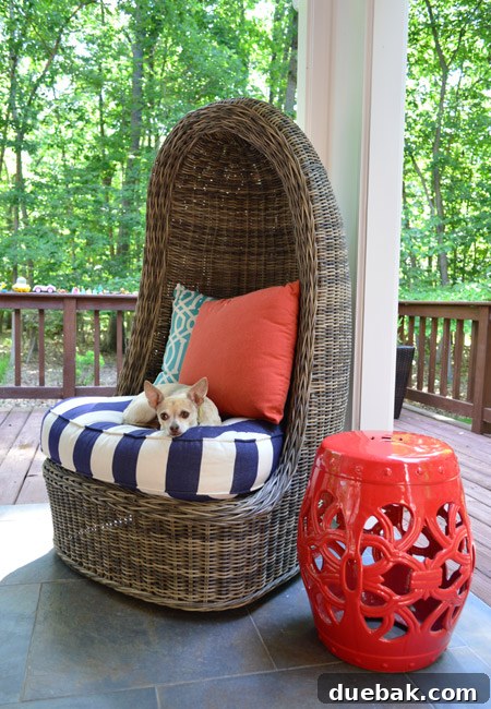 A vibrant red ceramic garden stool serving as a striking accent piece next to the comfortable wicker egg chair on a modern back porch