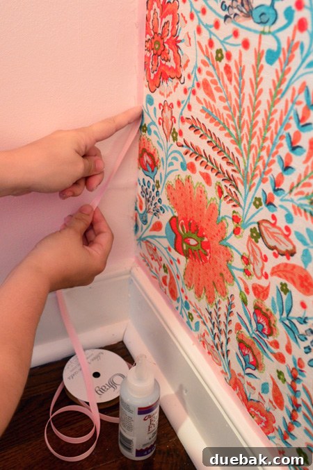 Transform Your Walls with Fabric 14 Pink ribbon being applied to cover the stapled edges of the fabric
