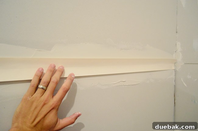 hand pushing drywall tape into bed of drywall mud along seam