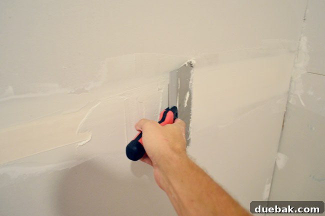 larger drywall knife being dragged across tape to apply another layer of mud
