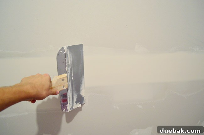 John dragging large drywall knife over seam in drywall to smooth mud