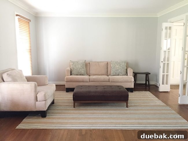Sparse, neutral living room before the makeover, featuring a light gray sofa, armchair, rug, and tufted ottoman, lacking decorative elements.