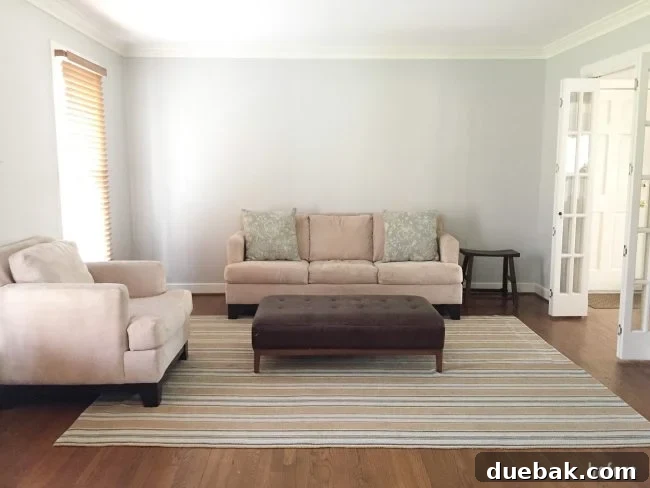 Before shot of the neutral living room, showing sparse furniture arrangement with a sofa, armchair, rug, and ottoman.