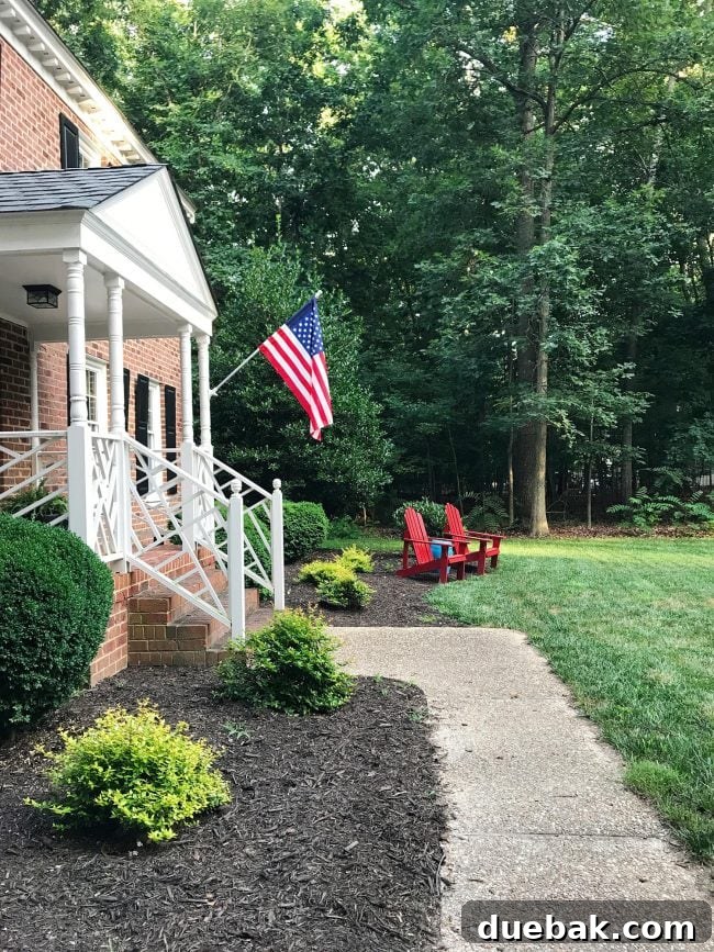 Patriotic Curb Appeal 2 A charming patriotic front porch featuring an American flag, classic red Adirondack chairs, and a vibrant blue garden stool, set against a welcoming colonial home facade.