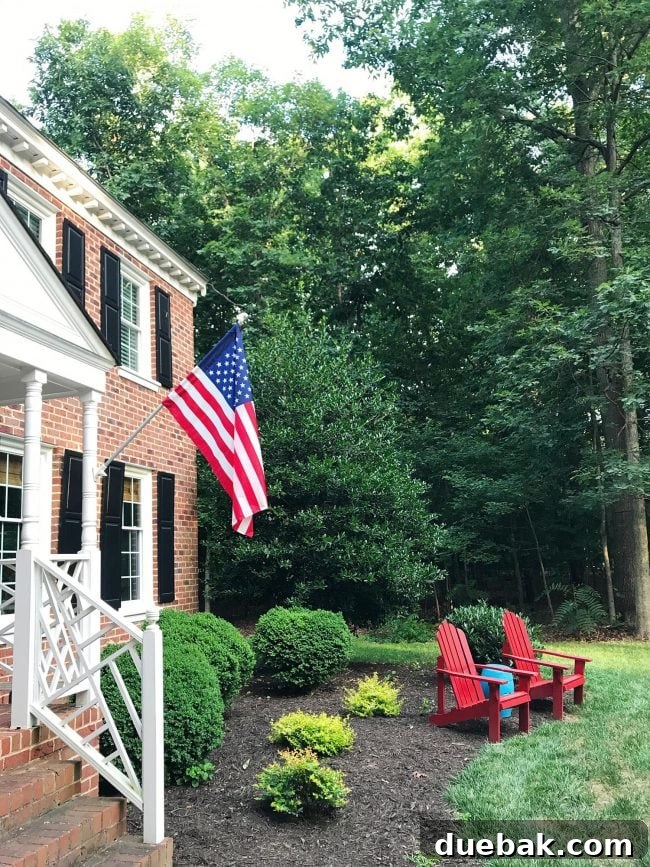 Patriotic Curb Appeal 8 An American flag proudly displayed on a brick colonial home, complemented by comfortable red Adirondack chairs on the porch, creating an inviting outdoor scene.
