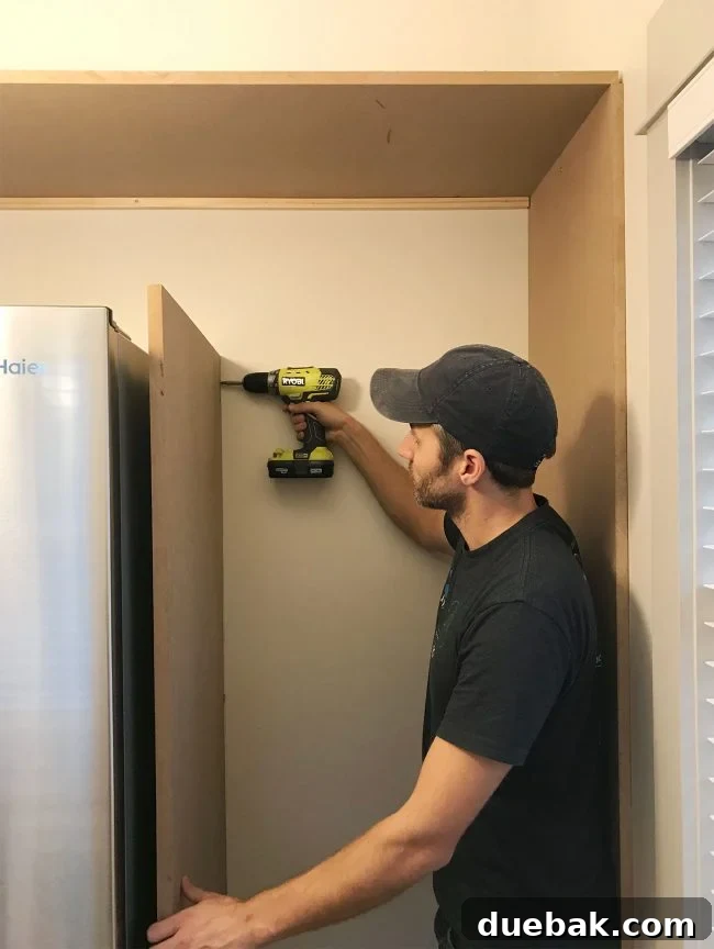 John drilling vertical MDF panel into wood already placed near fridge in pantry