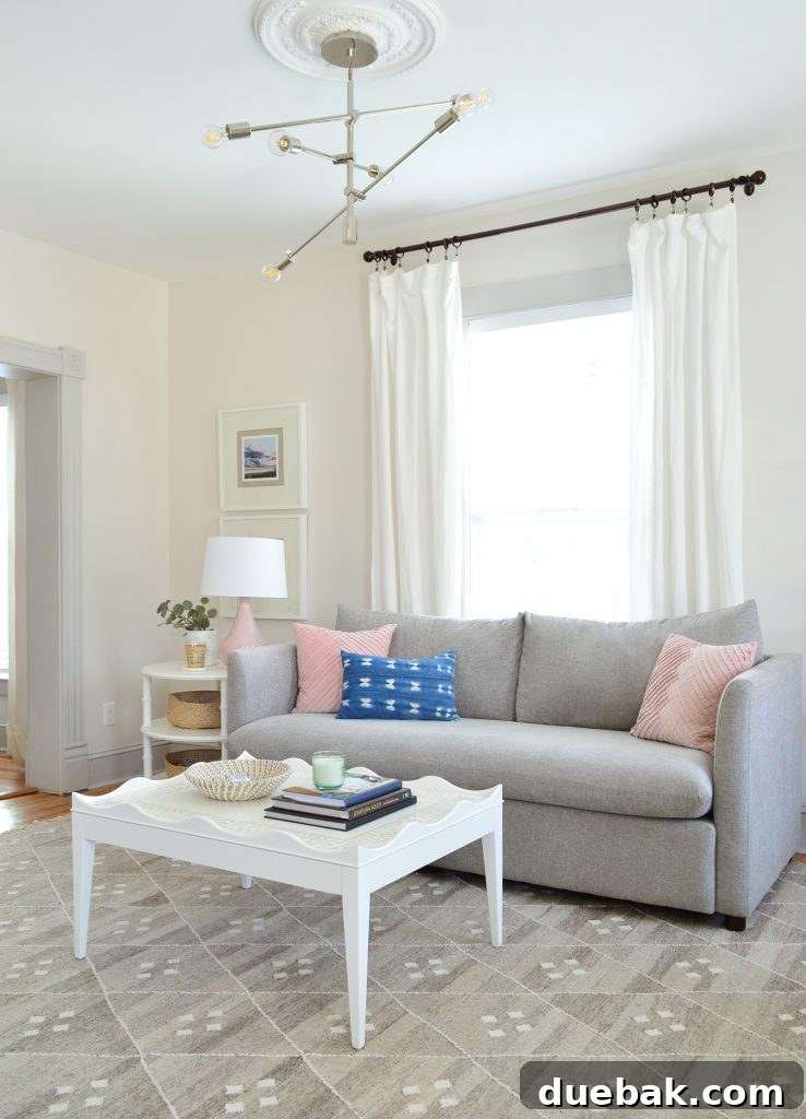 Bright neutral living room featuring a gray performance fabric sofa, white scalloped coffee table, and a modern chandelier, exuding beach house elegance.