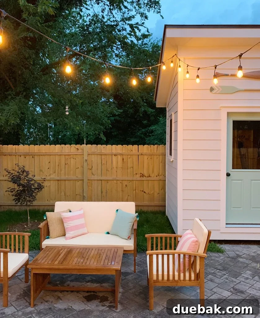 Cozy duplex backyard at dusk with glowing string lights, comfortable outdoor seating, a charming shed with decorative oars, and a back door painted in Pale Patina by Sherwin Williams.