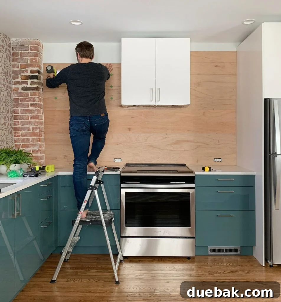John Standing On Ladder Nailing Wood Paneled Backsplash In Blue Kitchen