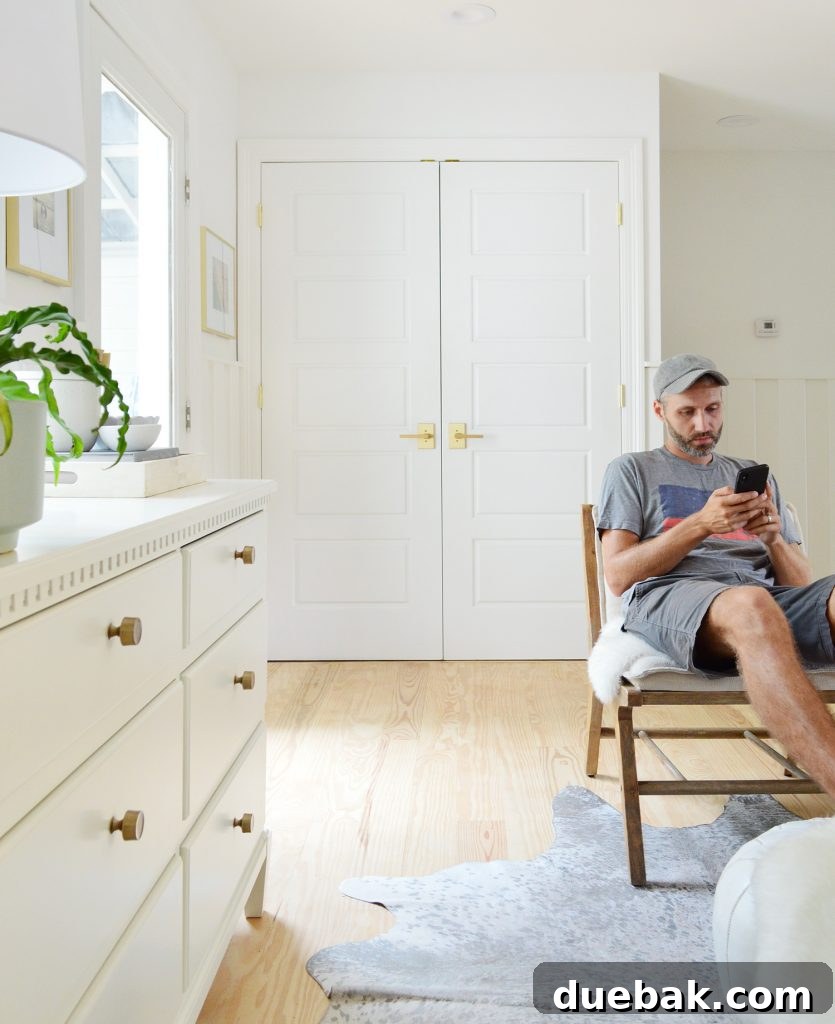 Compact Kitchen Living: Three Weeks In 17 John sits on a chair in a casual sitting area, looking at his phone, offering a perspective of an adult in the space. A dresser with gold hardware and a patterned rug are also visible, hinting at the home's cohesive design.