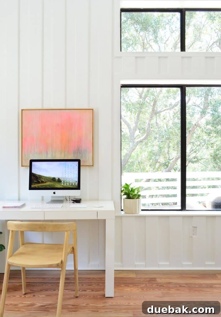 Life in Our Upstairs Family Room Four Weeks In 17 Adult workspace in a beachy living room, featuring a parsons desk, a comfortable office chair, and views of lush greenery through surrounding windows, with planked white walls, fostering a productive and inspiring environment.