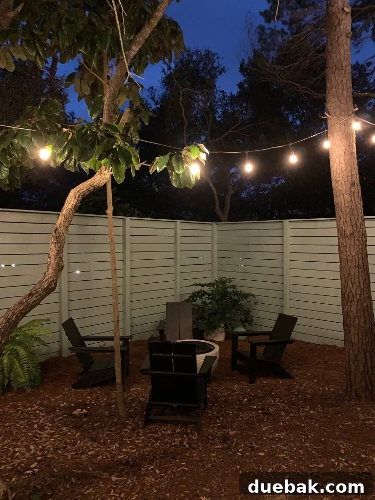 Nighttime view of fenced side yard with black Adirondack chairs around fire pit