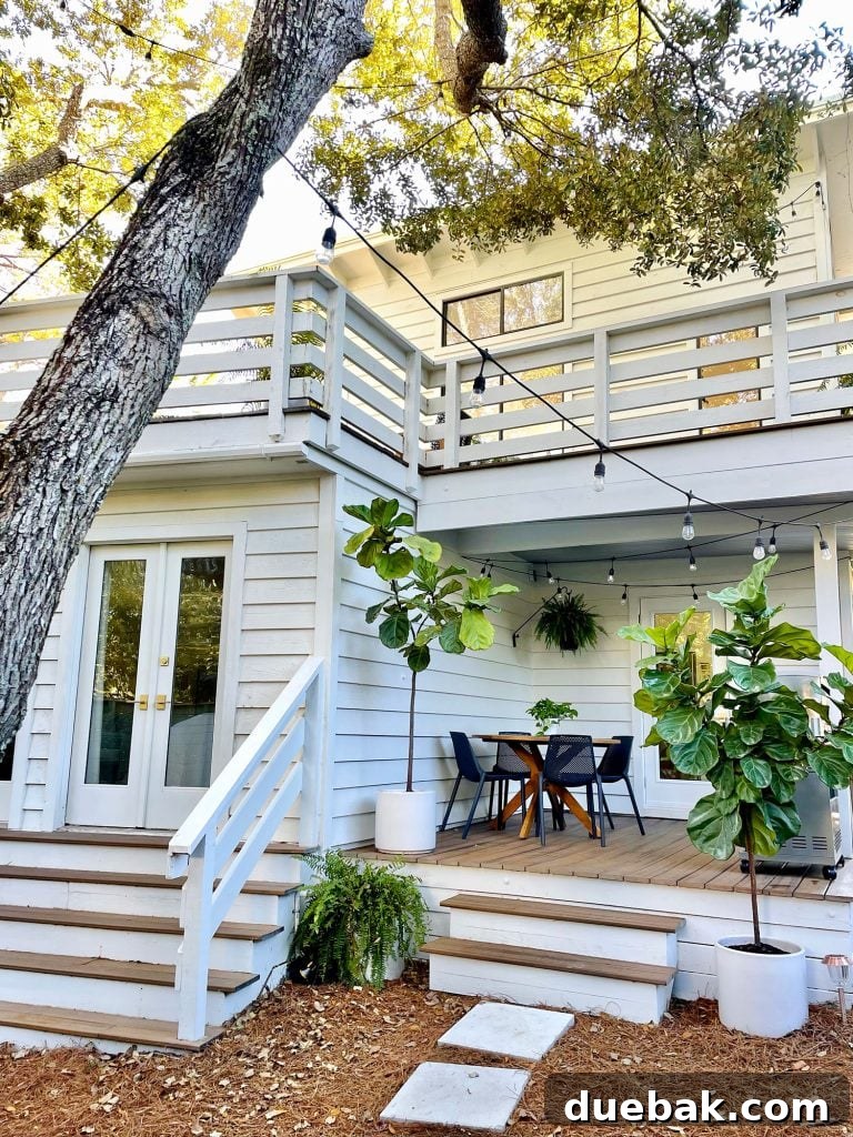 A picturesque view of the side porch, featuring a majestic oak tree in the foreground, against the backdrop of a white house adorned with string lights and thriving fig trees